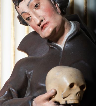 Zdjęcie nr 8: Rzeźba pełnoplastyczna, przyścienna ukazująca jezuickiego świętego św. Franciszka Borgiasza w całej postaci, w ujęciu frontalnym. Święty o szczupłej sylwetce stoi w kontrapoście, z lewą nogą ugiętą w kolanie i wysuniętą stopą, z głową przechyloną na prawy bark. Lewa ręka opuszczona wzdłuż tułowia, w prawej ugiętej w łokciu trzyma na wysokości piersi czaszkę ludzką. Twarz szczupła, o jasnej karnacji, z długim nosem i wzrokiem skierowanym d dół; włosy krótkie, zmierzwione. Ubrany w czarną sutannę jezuicką i płaszcz. 