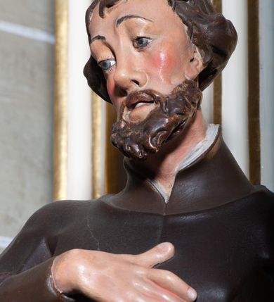 Zdjęcie nr 8: Rzeźba pełnoplastyczna, przyścienna, ukazująca jezuickiego świętego św. Franciszka Ksawerego w całej postaci, w ujęciu frontalnym. Święty o szczupłej sylwetce stoi w kontrapoście, z lewą nogą ugiętą w kolanie i wysuniętą lewą stopą, z głową przechyloną na prawy bark. Lewa ręka ugięta w łokciu z dłonią wysuniętą do przodu; prawa dłoń złożona na piersi. Twarz pociągła, z długim nosem, wydatnymi kośćmi policzkowymi i wzrokiem skierowanym d dół, okolona krótkimi włosami i krótkim ciemnym zarostem. Karnacja jasna z zaróżowionymi policzkami. Ubrany w czarną sutannę jezuicką z czarną pelerynką.