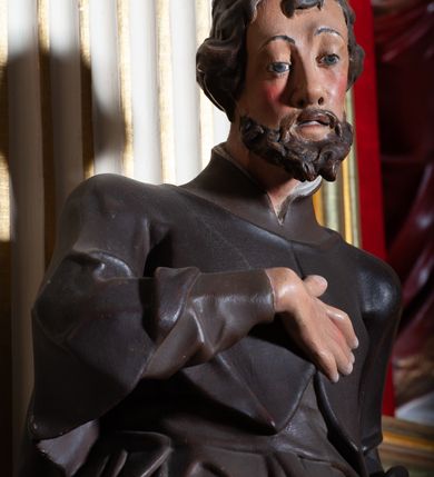 Zdjęcie nr 7: Rzeźba pełnoplastyczna, przyścienna, ukazująca jezuickiego świętego św. Franciszka Ksawerego w całej postaci, w ujęciu frontalnym. Święty o szczupłej sylwetce stoi w kontrapoście, z lewą nogą ugiętą w kolanie i wysuniętą lewą stopą, z głową przechyloną na prawy bark. Lewa ręka ugięta w łokciu z dłonią wysuniętą do przodu; prawa dłoń złożona na piersi. Twarz pociągła, z długim nosem, wydatnymi kośćmi policzkowymi i wzrokiem skierowanym d dół, okolona krótkimi włosami i krótkim ciemnym zarostem. Karnacja jasna z zaróżowionymi policzkami. Ubrany w czarną sutannę jezuicką z czarną pelerynką.