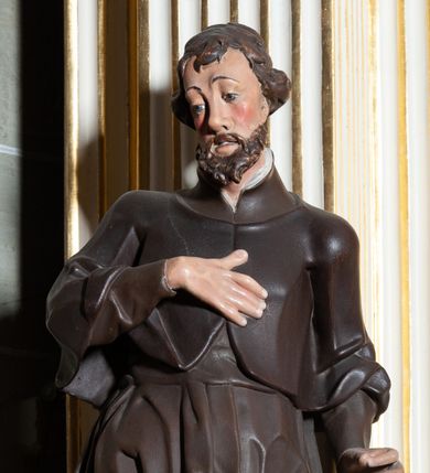 Zdjęcie nr 1: Rzeźba pełnoplastyczna, przyścienna, ukazująca jezuickiego świętego św. Franciszka Ksawerego w całej postaci, w ujęciu frontalnym. Święty o szczupłej sylwetce stoi w kontrapoście, z lewą nogą ugiętą w kolanie i wysuniętą lewą stopą, z głową przechyloną na prawy bark. Lewa ręka ugięta w łokciu z dłonią wysuniętą do przodu; prawa dłoń złożona na piersi. Twarz pociągła, z długim nosem, wydatnymi kośćmi policzkowymi i wzrokiem skierowanym d dół, okolona krótkimi włosami i krótkim ciemnym zarostem. Karnacja jasna z zaróżowionymi policzkami. Ubrany w czarną sutannę jezuicką z czarną pelerynką.