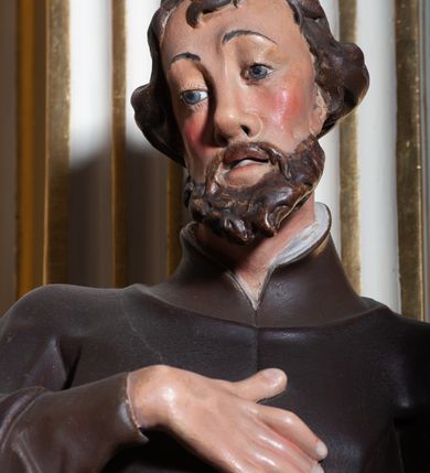 Zdjęcie nr 4: Rzeźba pełnoplastyczna, przyścienna, ukazująca jezuickiego świętego św. Franciszka Ksawerego w całej postaci, w ujęciu frontalnym. Święty o szczupłej sylwetce stoi w kontrapoście, z lewą nogą ugiętą w kolanie i wysuniętą lewą stopą, z głową przechyloną na prawy bark. Lewa ręka ugięta w łokciu z dłonią wysuniętą do przodu; prawa dłoń złożona na piersi. Twarz pociągła, z długim nosem, wydatnymi kośćmi policzkowymi i wzrokiem skierowanym d dół, okolona krótkimi włosami i krótkim ciemnym zarostem. Karnacja jasna z zaróżowionymi policzkami. Ubrany w czarną sutannę jezuicką z czarną pelerynką.