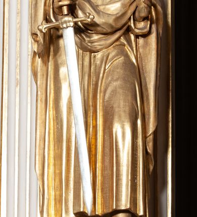 Zdjęcie nr 2: Rzeźba pełnoplastyczna, przyścienna, ustawiona na prostopadłościennym, wykonanym z czarnego marmuru relikwiarzu różnych świętych. Święty przedstawiony w całej postaci, stoi w lekkim kontrapoście, z rękami ugiętymi w łokciach, prawą dłonią trzyma rękojeść długiego srebrzystego miecza. Twarz pociągła okolona kędzierzawymi siwymi włosami i długą brodą. Ubrany w długą, sięgającą do kostek złotą  suknię i zarzucony na ramiona złoty płaszcz. 