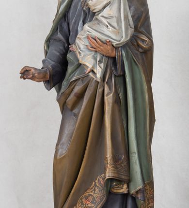 Zdjęcie nr 8: Feretron z rzeźbą św. Józefa z Dzieciątkiem ustawiony na wysokim postumencie. Postument zwężający się ku górze,  po  bokach flankowany dwiema wolutami, zaopatrzony w otwory na drążki do noszenia, stojący na czterech lwich łapkach. Na ściance frontowej zdobiony esownicami utworzonymi z liści akantu, kratką pośrodku oraz muszlą palmetową powyżej. Rzeźba ustawiona na niskim, kwadratowym, ciemnozielonym, cokole o ściętych narożach. Święty Józef ukazany w postawie stojącej, w delikatnym kontrapoście z Dzieciątkiem Jezus na lewym ręku, z prawą ręką wysuniętą przed siebie. Twarz podłużna, z długim i wąskim nosem, okolona krótko przystrzyżoną brodą; włosy brązowe, średniej długości, kręcone. Święty jest ubrany jest w ciemnofioletową suknię z długimi rękawami i ze złotą lamówką u dołu oraz przy szyi, przepasaną złotym paskiem; jasnobrązowy płaszcz z zieloną podszewką na brzegu zdobiony wicią roślinną  z owocami granatu; stopy bose. Dzieciątko Jezus ukazane w pozycji siedzącej na lewym ramieniu św. Józefa, prawą rączką obejmuje go za szyję, lewą kładzie na jego piersi. Twarz okrągła, wysokie czoło, małe usta, jasnobłękitne oczy, nos krótki i szeroki; włosy krótkie, jasne i kręcone. Dzieciątko jest ubrane w białą, długą sukienkę  z długimi rękawami, ze złotym obszyciem na brzegach, zdobioną malowanymi, złotymi małymi krzyżykami. Struktura postumentu polichromowana w kolorze jasnoszarym, detale  i ornamentyka złocone.
