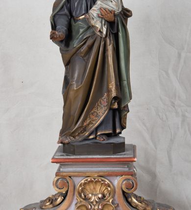 Zdjęcie nr 4: Feretron z rzeźbą św. Józefa z Dzieciątkiem ustawiony na wysokim postumencie. Postument zwężający się ku górze,  po  bokach flankowany dwiema wolutami, zaopatrzony w otwory na drążki do noszenia, stojący na czterech lwich łapkach. Na ściance frontowej zdobiony esownicami utworzonymi z liści akantu, kratką pośrodku oraz muszlą palmetową powyżej. Rzeźba ustawiona na niskim, kwadratowym, ciemnozielonym, cokole o ściętych narożach. Święty Józef ukazany w postawie stojącej, w delikatnym kontrapoście z Dzieciątkiem Jezus na lewym ręku, z prawą ręką wysuniętą przed siebie. Twarz podłużna, z długim i wąskim nosem, okolona krótko przystrzyżoną brodą; włosy brązowe, średniej długości, kręcone. Święty jest ubrany jest w ciemnofioletową suknię z długimi rękawami i ze złotą lamówką u dołu oraz przy szyi, przepasaną złotym paskiem; jasnobrązowy płaszcz z zieloną podszewką na brzegu zdobiony wicią roślinną  z owocami granatu; stopy bose. Dzieciątko Jezus ukazane w pozycji siedzącej na lewym ramieniu św. Józefa, prawą rączką obejmuje go za szyję, lewą kładzie na jego piersi. Twarz okrągła, wysokie czoło, małe usta, jasnobłękitne oczy, nos krótki i szeroki; włosy krótkie, jasne i kręcone. Dzieciątko jest ubrane w białą, długą sukienkę  z długimi rękawami, ze złotym obszyciem na brzegach, zdobioną malowanymi, złotymi małymi krzyżykami. Struktura postumentu polichromowana w kolorze jasnoszarym, detale  i ornamentyka złocone.
