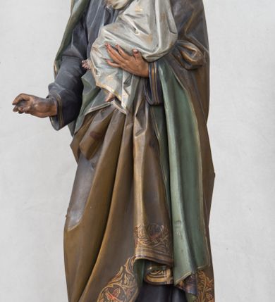 Zdjęcie nr 10: Feretron z rzeźbą św. Józefa z Dzieciątkiem ustawiony na wysokim postumencie. Postument zwężający się ku górze,  po  bokach flankowany dwiema wolutami, zaopatrzony w otwory na drążki do noszenia, stojący na czterech lwich łapkach. Na ściance frontowej zdobiony esownicami utworzonymi z liści akantu, kratką pośrodku oraz muszlą palmetową powyżej. Rzeźba ustawiona na niskim, kwadratowym, ciemnozielonym, cokole o ściętych narożach. Święty Józef ukazany w postawie stojącej, w delikatnym kontrapoście z Dzieciątkiem Jezus na lewym ręku, z prawą ręką wysuniętą przed siebie. Twarz podłużna, z długim i wąskim nosem, okolona krótko przystrzyżoną brodą; włosy brązowe, średniej długości, kręcone. Święty jest ubrany jest w ciemnofioletową suknię z długimi rękawami i ze złotą lamówką u dołu oraz przy szyi, przepasaną złotym paskiem; jasnobrązowy płaszcz z zieloną podszewką na brzegu zdobiony wicią roślinną  z owocami granatu; stopy bose. Dzieciątko Jezus ukazane w pozycji siedzącej na lewym ramieniu św. Józefa, prawą rączką obejmuje go za szyję, lewą kładzie na jego piersi. Twarz okrągła, wysokie czoło, małe usta, jasnobłękitne oczy, nos krótki i szeroki; włosy krótkie, jasne i kręcone. Dzieciątko jest ubrane w białą, długą sukienkę  z długimi rękawami, ze złotym obszyciem na brzegach, zdobioną malowanymi, złotymi małymi krzyżykami. Struktura postumentu polichromowana w kolorze jasnoszarym, detale  i ornamentyka złocone.
