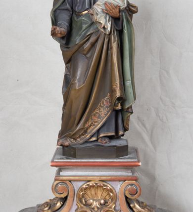 Zdjęcie nr 5: Feretron z rzeźbą św. Józefa z Dzieciątkiem ustawiony na wysokim postumencie. Postument zwężający się ku górze,  po  bokach flankowany dwiema wolutami, zaopatrzony w otwory na drążki do noszenia, stojący na czterech lwich łapkach. Na ściance frontowej zdobiony esownicami utworzonymi z liści akantu, kratką pośrodku oraz muszlą palmetową powyżej. Rzeźba ustawiona na niskim, kwadratowym, ciemnozielonym, cokole o ściętych narożach. Święty Józef ukazany w postawie stojącej, w delikatnym kontrapoście z Dzieciątkiem Jezus na lewym ręku, z prawą ręką wysuniętą przed siebie. Twarz podłużna, z długim i wąskim nosem, okolona krótko przystrzyżoną brodą; włosy brązowe, średniej długości, kręcone. Święty jest ubrany jest w ciemnofioletową suknię z długimi rękawami i ze złotą lamówką u dołu oraz przy szyi, przepasaną złotym paskiem; jasnobrązowy płaszcz z zieloną podszewką na brzegu zdobiony wicią roślinną  z owocami granatu; stopy bose. Dzieciątko Jezus ukazane w pozycji siedzącej na lewym ramieniu św. Józefa, prawą rączką obejmuje go za szyję, lewą kładzie na jego piersi. Twarz okrągła, wysokie czoło, małe usta, jasnobłękitne oczy, nos krótki i szeroki; włosy krótkie, jasne i kręcone. Dzieciątko jest ubrane w białą, długą sukienkę  z długimi rękawami, ze złotym obszyciem na brzegach, zdobioną malowanymi, złotymi małymi krzyżykami. Struktura postumentu polichromowana w kolorze jasnoszarym, detale  i ornamentyka złocone.
