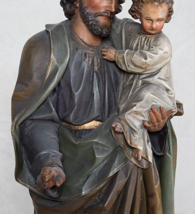 Zdjęcie nr 11: Feretron z rzeźbą św. Józefa z Dzieciątkiem ustawiony na wysokim postumencie. Postument zwężający się ku górze,  po  bokach flankowany dwiema wolutami, zaopatrzony w otwory na drążki do noszenia, stojący na czterech lwich łapkach. Na ściance frontowej zdobiony esownicami utworzonymi z liści akantu, kratką pośrodku oraz muszlą palmetową powyżej. Rzeźba ustawiona na niskim, kwadratowym, ciemnozielonym, cokole o ściętych narożach. Święty Józef ukazany w postawie stojącej, w delikatnym kontrapoście z Dzieciątkiem Jezus na lewym ręku, z prawą ręką wysuniętą przed siebie. Twarz podłużna, z długim i wąskim nosem, okolona krótko przystrzyżoną brodą; włosy brązowe, średniej długości, kręcone. Święty jest ubrany jest w ciemnofioletową suknię z długimi rękawami i ze złotą lamówką u dołu oraz przy szyi, przepasaną złotym paskiem; jasnobrązowy płaszcz z zieloną podszewką na brzegu zdobiony wicią roślinną  z owocami granatu; stopy bose. Dzieciątko Jezus ukazane w pozycji siedzącej na lewym ramieniu św. Józefa, prawą rączką obejmuje go za szyję, lewą kładzie na jego piersi. Twarz okrągła, wysokie czoło, małe usta, jasnobłękitne oczy, nos krótki i szeroki; włosy krótkie, jasne i kręcone. Dzieciątko jest ubrane w białą, długą sukienkę  z długimi rękawami, ze złotym obszyciem na brzegach, zdobioną malowanymi, złotymi małymi krzyżykami. Struktura postumentu polichromowana w kolorze jasnoszarym, detale  i ornamentyka złocone.
