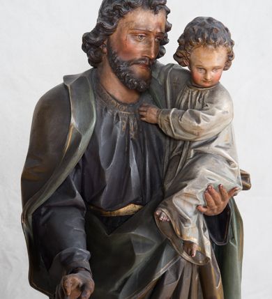 Zdjęcie nr 3: Feretron z rzeźbą św. Józefa z Dzieciątkiem ustawiony na wysokim postumencie. Postument zwężający się ku górze,  po  bokach flankowany dwiema wolutami, zaopatrzony w otwory na drążki do noszenia, stojący na czterech lwich łapkach. Na ściance frontowej zdobiony esownicami utworzonymi z liści akantu, kratką pośrodku oraz muszlą palmetową powyżej. Rzeźba ustawiona na niskim, kwadratowym, ciemnozielonym, cokole o ściętych narożach. Święty Józef ukazany w postawie stojącej, w delikatnym kontrapoście z Dzieciątkiem Jezus na lewym ręku, z prawą ręką wysuniętą przed siebie. Twarz podłużna, z długim i wąskim nosem, okolona krótko przystrzyżoną brodą; włosy brązowe, średniej długości, kręcone. Święty jest ubrany jest w ciemnofioletową suknię z długimi rękawami i ze złotą lamówką u dołu oraz przy szyi, przepasaną złotym paskiem; jasnobrązowy płaszcz z zieloną podszewką na brzegu zdobiony wicią roślinną  z owocami granatu; stopy bose. Dzieciątko Jezus ukazane w pozycji siedzącej na lewym ramieniu św. Józefa, prawą rączką obejmuje go za szyję, lewą kładzie na jego piersi. Twarz okrągła, wysokie czoło, małe usta, jasnobłękitne oczy, nos krótki i szeroki; włosy krótkie, jasne i kręcone. Dzieciątko jest ubrane w białą, długą sukienkę  z długimi rękawami, ze złotym obszyciem na brzegach, zdobioną malowanymi, złotymi małymi krzyżykami. Struktura postumentu polichromowana w kolorze jasnoszarym, detale  i ornamentyka złocone.
