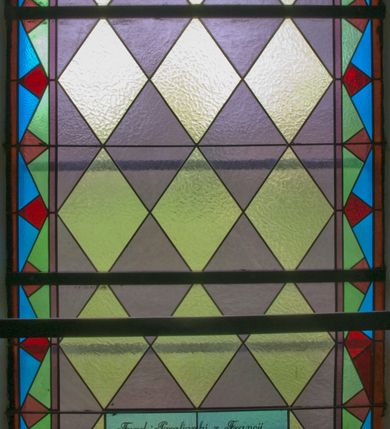 Zdjęcie nr 4: Witraż w kształcie stojącego prostokąta zamkniętego łukiem półkolistym ujęty geometryczną bordiurą wypełnioną motywem białych i niebieskich trójkątów oraz ciemnoczerwonych rombów. W polu ukośna kratka wypełniona ułożonymi naprzemiennie białymi i jasnofioletowymi rombami. W dolnej części witraża owalny medalion wsparty na girlandzie białych, kwitnących róż mieszczący popiersie Matki Boskiej z Dzieciątkiem ukazanej w typie Matki Boskiej Nieustającej Pomocy umieszczone na złotym tle. Maria zwrócona w trzech czwartych w lewo, na lewym przedramieniu trzyma siedzące Dzieciątko, prawą dłonią podtrzymuje dłonie Jezusa. Matka Boska ubrana jest w czerwoną suknię z długimi rękawami wykończoną przy dekolcie złotą lamówką oraz niebieski płaszcz zarzucony na głowę. Dzieciątko jest zwrócone w trzech czwartych w prawo z głową w przeciwnym kierunku. Jezus nosi jasnozieloną tunikę z długimi, szerokimi rękawami oraz złoty, szrafowany płaszcz, spod którego widoczne są bose stopy. Wokół głowy Marii znajduje się biały nimb wypełniony bordiurą roślinną, wokół głowy Dzieciątka nimb krzyżowy. Obie postaci ukazane są w złotych, kameryzowanych koronach typu otwartego. Powyżej medalionu prostokątne pole z napisem: „Fund[acja] Parafianki z Francji / Wykonano R[oku] P[ańskiego] 1939 za pobytu Ks[iędza] Prob[oszcza] Wł[adysława] Ziemińskiego”. 