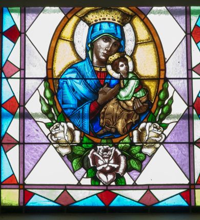 Zdjęcie nr 2: Witraż w kształcie stojącego prostokąta zamkniętego łukiem półkolistym ujęty geometryczną bordiurą wypełnioną motywem białych i niebieskich trójkątów oraz ciemnoczerwonych rombów. W polu ukośna kratka wypełniona ułożonymi naprzemiennie białymi i jasnofioletowymi rombami. W dolnej części witraża owalny medalion wsparty na girlandzie białych, kwitnących róż mieszczący popiersie Matki Boskiej z Dzieciątkiem ukazanej w typie Matki Boskiej Nieustającej Pomocy umieszczone na złotym tle. Maria zwrócona w trzech czwartych w lewo, na lewym przedramieniu trzyma siedzące Dzieciątko, prawą dłonią podtrzymuje dłonie Jezusa. Matka Boska ubrana jest w czerwoną suknię z długimi rękawami wykończoną przy dekolcie złotą lamówką oraz niebieski płaszcz zarzucony na głowę. Dzieciątko jest zwrócone w trzech czwartych w prawo z głową w przeciwnym kierunku. Jezus nosi jasnozieloną tunikę z długimi, szerokimi rękawami oraz złoty, szrafowany płaszcz, spod którego widoczne są bose stopy. Wokół głowy Marii znajduje się biały nimb wypełniony bordiurą roślinną, wokół głowy Dzieciątka nimb krzyżowy. Obie postaci ukazane są w złotych, kameryzowanych koronach typu otwartego. Powyżej medalionu prostokątne pole z napisem: „Fund[acja] Parafianki z Francji / Wykonano R[oku] P[ańskiego] 1939 za pobytu Ks[iędza] Prob[oszcza] Wł[adysława] Ziemińskiego”. 