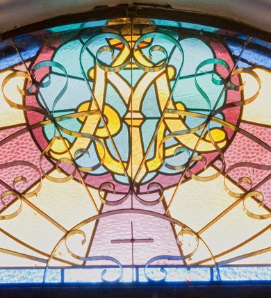 Zdjęcie nr 1: Witraż w kształcie odcinka koła. W górnej części kompozycji umieszczony okrągły medalion ze złotym monogramem maryjnym umieszczonym na jasnobłękitnym polu w kształcie wieloliścia ujętego bordową ramą. W tle naprzemiennie ułożone promienie w kolorach jasnożółtym i fioletowym. Całość ujęta prostą, niebieską ramą.


