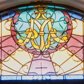 Zdjęcie nr 1: Witraż w kształcie odcinka koła. W górnej części kompozycji umieszczony okrągły medalion ze złotym monogramem maryjnym umieszczonym na jasnobłękitnym polu w kształcie wieloliścia ujętego bordową ramą. W tle naprzemiennie ułożone promienie w kolorach jasnożółtym i fioletowym. Całość ujęta prostą, niebieską ramą.


