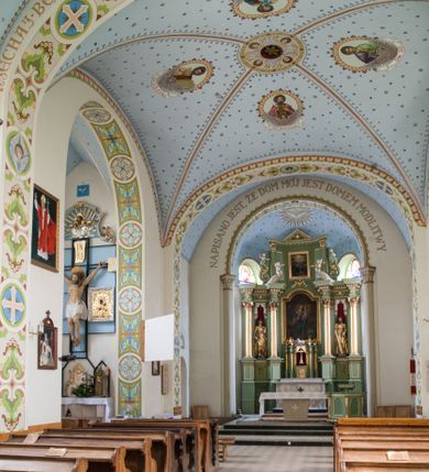Zdjęcie nr 2: W prezbiterium do wysokości parapetów okiennych ściany malowane jednolicie w kolorze beżowym. Powyżej, na sklepieniu motyw gwiaździstego nieba z niewielkimi malowanymi złotem gwiazdkami na jasnobłękitnym tle. Ponad ołtarzem głównym gołębica Ducha Świętego na tle glorii promienistej tworzącej motyw dekoracyjnej rozety. Obramienia okienne zaznaczone geometrycznymi, złotymi lamówkami; rozglifienia okien białe. W podniebiu arkady tęczowej pas stylizowanego, symetrycznego ornamentu roślinnego w kolorach jasnoniebieskim, beżowym i jasnobrązowym. W osi uskrzydlona główka anielska. Na licu ściany tęczowej majuskułowy, złoty napis „NAPISANO JEST, ŻE DOM MÓJ JEST DOMEM MODLITWY”. W nawie głównej i transepcie ściany malowane jednolicie w kolorze beżowym. W południowym ramieniu transeptu, ponad ołtarzem motyw czteroliścia z wpisanym medalionem mieszczącym gorejące serce Jezusa, oplecione cierniową koroną i zwieńczone krzyżem. Na ścianie zachodniej, ponad chórem muzycznym dwie ustawione przeciwstawnie grupy muzykujących aniołów umieszczone na tle obłoków oraz majuskułowy, złoty napis „ŚPIEWAJCIE PANU W RADOŚCI”. Na artykułujących nawę filarach przechodzących w gurty sklepienne stylizowany, symetryczny ornament roślinny o przebiegu pasowym z wplecionymi motywami rozet z wpisanymi krzyżami równoramiennymi lub uskrzydlonymi główkami cherubinów. Ornament utrzymany w kolorach jasnozielonym, błękitnym, jasnobrązowym i beżowym. Na licu gurtu sklepiennego od strony zachodniej majuskułowy, złoty napis „NIECHAJ BĘDZIE POCHWALONY PRZENAJŚWIĘTSZY SAKRAMENT”. Sklepienie krzyżowe na przecięciu naw i ramionach transeptu oraz kolebkowe w nawie głównej malowane kolorem jasnobłękitnym w motyw złoconych gwiazd i rozetek. W wysklepkach, na przecięciu naw medaliony z popiersiami czterech ewangelistów. Od strony prezbiterium św. Mateusz ukazany jako dojrzały, ciemnowłosy mężczyzna piszący piórem w otwartej księdze, ubrany w czerwono-zielone szaty. Poniżej anioł – symbol św. Mateusza oraz banderola z napisem „ŚW(IĘTY) MATEUSZ”. Od południa medalion z popiersiem św. Jana ukazanego jako jasnowłosy młodzieniec, ubrany w zielono-fioletowe szaty, trzymający w ręku kielich. Poniżej orzeł o szeroko rozpostartych skrzydłach – symbol św. Jana oraz banderola z napisem „ŚW(IĘTY) JAN”. Od strony zachodniej medalion mieszczący popiersie św. Marka przedstawionego jako dojrzały mężczyzna o krótkich, ciemnych włosach i brodzie ubrany w żółto-niebieskie szaty. Święty trzyma w ręku zamkniętą księgę. Poniżej uskrzydlony lew – symbol św. Marka oraz banderola z napisem „ŚW(IĘTY) MAREK”. Od północy medalion z popiersiem św. Łukasza ukazanego jako dojrzały mężczyzna o krótkich, ciemnych włosach i brodzie trzymający w ręku obraz oraz laskę. Poniżej uskrzydlony byk oraz banderola z napisem „ŚW(IĘTY) ŁUKASZ”. W części centralnej tego przęsła medalion z czterema uskrzydlonymi główkami anielskimi. W centrum kolebkowego sklepienia ponad nawą główną pole w kształcie stojącego prostokąta zamkniętego łukiem półkolistym mieszczące przedstawienie Matki Boskiej Niepokalanie Poczętej na tle obłoków oraz aniołów i uskrzydlonych główek anielskich. Maria została przedstawiona jako młoda, jasnowłosa kobieta z rękami skrzyżowanymi na piersi, ubrana w jasnoróżową suknię oraz błękitny płaszcz. Pod jej stopami ukazano sierp księżyca. 