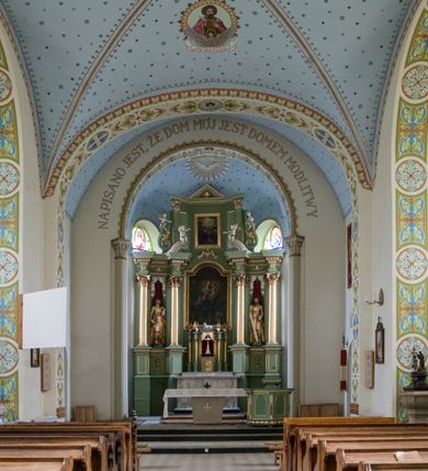 Zdjęcie nr 5: W prezbiterium do wysokości parapetów okiennych ściany malowane jednolicie w kolorze beżowym. Powyżej, na sklepieniu motyw gwiaździstego nieba z niewielkimi malowanymi złotem gwiazdkami na jasnobłękitnym tle. Ponad ołtarzem głównym gołębica Ducha Świętego na tle glorii promienistej tworzącej motyw dekoracyjnej rozety. Obramienia okienne zaznaczone geometrycznymi, złotymi lamówkami; rozglifienia okien białe. W podniebiu arkady tęczowej pas stylizowanego, symetrycznego ornamentu roślinnego w kolorach jasnoniebieskim, beżowym i jasnobrązowym. W osi uskrzydlona główka anielska. Na licu ściany tęczowej majuskułowy, złoty napis „NAPISANO JEST, ŻE DOM MÓJ JEST DOMEM MODLITWY”. W nawie głównej i transepcie ściany malowane jednolicie w kolorze beżowym. W południowym ramieniu transeptu, ponad ołtarzem motyw czteroliścia z wpisanym medalionem mieszczącym gorejące serce Jezusa, oplecione cierniową koroną i zwieńczone krzyżem. Na ścianie zachodniej, ponad chórem muzycznym dwie ustawione przeciwstawnie grupy muzykujących aniołów umieszczone na tle obłoków oraz majuskułowy, złoty napis „ŚPIEWAJCIE PANU W RADOŚCI”. Na artykułujących nawę filarach przechodzących w gurty sklepienne stylizowany, symetryczny ornament roślinny o przebiegu pasowym z wplecionymi motywami rozet z wpisanymi krzyżami równoramiennymi lub uskrzydlonymi główkami cherubinów. Ornament utrzymany w kolorach jasnozielonym, błękitnym, jasnobrązowym i beżowym. Na licu gurtu sklepiennego od strony zachodniej majuskułowy, złoty napis „NIECHAJ BĘDZIE POCHWALONY PRZENAJŚWIĘTSZY SAKRAMENT”. Sklepienie krzyżowe na przecięciu naw i ramionach transeptu oraz kolebkowe w nawie głównej malowane kolorem jasnobłękitnym w motyw złoconych gwiazd i rozetek. W wysklepkach, na przecięciu naw medaliony z popiersiami czterech ewangelistów. Od strony prezbiterium św. Mateusz ukazany jako dojrzały, ciemnowłosy mężczyzna piszący piórem w otwartej księdze, ubrany w czerwono-zielone szaty. Poniżej anioł – symbol św. Mateusza oraz banderola z napisem „ŚW(IĘTY) MATEUSZ”. Od południa medalion z popiersiem św. Jana ukazanego jako jasnowłosy młodzieniec, ubrany w zielono-fioletowe szaty, trzymający w ręku kielich. Poniżej orzeł o szeroko rozpostartych skrzydłach – symbol św. Jana oraz banderola z napisem „ŚW(IĘTY) JAN”. Od strony zachodniej medalion mieszczący popiersie św. Marka przedstawionego jako dojrzały mężczyzna o krótkich, ciemnych włosach i brodzie ubrany w żółto-niebieskie szaty. Święty trzyma w ręku zamkniętą księgę. Poniżej uskrzydlony lew – symbol św. Marka oraz banderola z napisem „ŚW(IĘTY) MAREK”. Od północy medalion z popiersiem św. Łukasza ukazanego jako dojrzały mężczyzna o krótkich, ciemnych włosach i brodzie trzymający w ręku obraz oraz laskę. Poniżej uskrzydlony byk oraz banderola z napisem „ŚW(IĘTY) ŁUKASZ”. W części centralnej tego przęsła medalion z czterema uskrzydlonymi główkami anielskimi. W centrum kolebkowego sklepienia ponad nawą główną pole w kształcie stojącego prostokąta zamkniętego łukiem półkolistym mieszczące przedstawienie Matki Boskiej Niepokalanie Poczętej na tle obłoków oraz aniołów i uskrzydlonych główek anielskich. Maria została przedstawiona jako młoda, jasnowłosa kobieta z rękami skrzyżowanymi na piersi, ubrana w jasnoróżową suknię oraz błękitny płaszcz. Pod jej stopami ukazano sierp księżyca. 