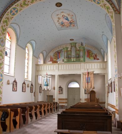 Zdjęcie nr 8: W prezbiterium do wysokości parapetów okiennych ściany malowane jednolicie w kolorze beżowym. Powyżej, na sklepieniu motyw gwiaździstego nieba z niewielkimi malowanymi złotem gwiazdkami na jasnobłękitnym tle. Ponad ołtarzem głównym gołębica Ducha Świętego na tle glorii promienistej tworzącej motyw dekoracyjnej rozety. Obramienia okienne zaznaczone geometrycznymi, złotymi lamówkami; rozglifienia okien białe. W podniebiu arkady tęczowej pas stylizowanego, symetrycznego ornamentu roślinnego w kolorach jasnoniebieskim, beżowym i jasnobrązowym. W osi uskrzydlona główka anielska. Na licu ściany tęczowej majuskułowy, złoty napis „NAPISANO JEST, ŻE DOM MÓJ JEST DOMEM MODLITWY”. W nawie głównej i transepcie ściany malowane jednolicie w kolorze beżowym. W południowym ramieniu transeptu, ponad ołtarzem motyw czteroliścia z wpisanym medalionem mieszczącym gorejące serce Jezusa, oplecione cierniową koroną i zwieńczone krzyżem. Na ścianie zachodniej, ponad chórem muzycznym dwie ustawione przeciwstawnie grupy muzykujących aniołów umieszczone na tle obłoków oraz majuskułowy, złoty napis „ŚPIEWAJCIE PANU W RADOŚCI”. Na artykułujących nawę filarach przechodzących w gurty sklepienne stylizowany, symetryczny ornament roślinny o przebiegu pasowym z wplecionymi motywami rozet z wpisanymi krzyżami równoramiennymi lub uskrzydlonymi główkami cherubinów. Ornament utrzymany w kolorach jasnozielonym, błękitnym, jasnobrązowym i beżowym. Na licu gurtu sklepiennego od strony zachodniej majuskułowy, złoty napis „NIECHAJ BĘDZIE POCHWALONY PRZENAJŚWIĘTSZY SAKRAMENT”. Sklepienie krzyżowe na przecięciu naw i ramionach transeptu oraz kolebkowe w nawie głównej malowane kolorem jasnobłękitnym w motyw złoconych gwiazd i rozetek. W wysklepkach, na przecięciu naw medaliony z popiersiami czterech ewangelistów. Od strony prezbiterium św. Mateusz ukazany jako dojrzały, ciemnowłosy mężczyzna piszący piórem w otwartej księdze, ubrany w czerwono-zielone szaty. Poniżej anioł – symbol św. Mateusza oraz banderola z napisem „ŚW(IĘTY) MATEUSZ”. Od południa medalion z popiersiem św. Jana ukazanego jako jasnowłosy młodzieniec, ubrany w zielono-fioletowe szaty, trzymający w ręku kielich. Poniżej orzeł o szeroko rozpostartych skrzydłach – symbol św. Jana oraz banderola z napisem „ŚW(IĘTY) JAN”. Od strony zachodniej medalion mieszczący popiersie św. Marka przedstawionego jako dojrzały mężczyzna o krótkich, ciemnych włosach i brodzie ubrany w żółto-niebieskie szaty. Święty trzyma w ręku zamkniętą księgę. Poniżej uskrzydlony lew – symbol św. Marka oraz banderola z napisem „ŚW(IĘTY) MAREK”. Od północy medalion z popiersiem św. Łukasza ukazanego jako dojrzały mężczyzna o krótkich, ciemnych włosach i brodzie trzymający w ręku obraz oraz laskę. Poniżej uskrzydlony byk oraz banderola z napisem „ŚW(IĘTY) ŁUKASZ”. W części centralnej tego przęsła medalion z czterema uskrzydlonymi główkami anielskimi. W centrum kolebkowego sklepienia ponad nawą główną pole w kształcie stojącego prostokąta zamkniętego łukiem półkolistym mieszczące przedstawienie Matki Boskiej Niepokalanie Poczętej na tle obłoków oraz aniołów i uskrzydlonych główek anielskich. Maria została przedstawiona jako młoda, jasnowłosa kobieta z rękami skrzyżowanymi na piersi, ubrana w jasnoróżową suknię oraz błękitny płaszcz. Pod jej stopami ukazano sierp księżyca. 