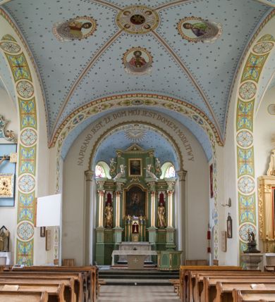 Zdjęcie nr 1: W prezbiterium do wysokości parapetów okiennych ściany malowane jednolicie w kolorze beżowym. Powyżej, na sklepieniu motyw gwiaździstego nieba z niewielkimi malowanymi złotem gwiazdkami na jasnobłękitnym tle. Ponad ołtarzem głównym gołębica Ducha Świętego na tle glorii promienistej tworzącej motyw dekoracyjnej rozety. Obramienia okienne zaznaczone geometrycznymi, złotymi lamówkami; rozglifienia okien białe. W podniebiu arkady tęczowej pas stylizowanego, symetrycznego ornamentu roślinnego w kolorach jasnoniebieskim, beżowym i jasnobrązowym. W osi uskrzydlona główka anielska. Na licu ściany tęczowej majuskułowy, złoty napis „NAPISANO JEST, ŻE DOM MÓJ JEST DOMEM MODLITWY”. W nawie głównej i transepcie ściany malowane jednolicie w kolorze beżowym. W południowym ramieniu transeptu, ponad ołtarzem motyw czteroliścia z wpisanym medalionem mieszczącym gorejące serce Jezusa, oplecione cierniową koroną i zwieńczone krzyżem. Na ścianie zachodniej, ponad chórem muzycznym dwie ustawione przeciwstawnie grupy muzykujących aniołów umieszczone na tle obłoków oraz majuskułowy, złoty napis „ŚPIEWAJCIE PANU W RADOŚCI”. Na artykułujących nawę filarach przechodzących w gurty sklepienne stylizowany, symetryczny ornament roślinny o przebiegu pasowym z wplecionymi motywami rozet z wpisanymi krzyżami równoramiennymi lub uskrzydlonymi główkami cherubinów. Ornament utrzymany w kolorach jasnozielonym, błękitnym, jasnobrązowym i beżowym. Na licu gurtu sklepiennego od strony zachodniej majuskułowy, złoty napis „NIECHAJ BĘDZIE POCHWALONY PRZENAJŚWIĘTSZY SAKRAMENT”. Sklepienie krzyżowe na przecięciu naw i ramionach transeptu oraz kolebkowe w nawie głównej malowane kolorem jasnobłękitnym w motyw złoconych gwiazd i rozetek. W wysklepkach, na przecięciu naw medaliony z popiersiami czterech ewangelistów. Od strony prezbiterium św. Mateusz ukazany jako dojrzały, ciemnowłosy mężczyzna piszący piórem w otwartej księdze, ubrany w czerwono-zielone szaty. Poniżej anioł – symbol św. Mateusza oraz banderola z napisem „ŚW(IĘTY) MATEUSZ”. Od południa medalion z popiersiem św. Jana ukazanego jako jasnowłosy młodzieniec, ubrany w zielono-fioletowe szaty, trzymający w ręku kielich. Poniżej orzeł o szeroko rozpostartych skrzydłach – symbol św. Jana oraz banderola z napisem „ŚW(IĘTY) JAN”. Od strony zachodniej medalion mieszczący popiersie św. Marka przedstawionego jako dojrzały mężczyzna o krótkich, ciemnych włosach i brodzie ubrany w żółto-niebieskie szaty. Święty trzyma w ręku zamkniętą księgę. Poniżej uskrzydlony lew – symbol św. Marka oraz banderola z napisem „ŚW(IĘTY) MAREK”. Od północy medalion z popiersiem św. Łukasza ukazanego jako dojrzały mężczyzna o krótkich, ciemnych włosach i brodzie trzymający w ręku obraz oraz laskę. Poniżej uskrzydlony byk oraz banderola z napisem „ŚW(IĘTY) ŁUKASZ”. W części centralnej tego przęsła medalion z czterema uskrzydlonymi główkami anielskimi. W centrum kolebkowego sklepienia ponad nawą główną pole w kształcie stojącego prostokąta zamkniętego łukiem półkolistym mieszczące przedstawienie Matki Boskiej Niepokalanie Poczętej na tle obłoków oraz aniołów i uskrzydlonych główek anielskich. Maria została przedstawiona jako młoda, jasnowłosa kobieta z rękami skrzyżowanymi na piersi, ubrana w jasnoróżową suknię oraz błękitny płaszcz. Pod jej stopami ukazano sierp księżyca. 