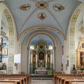 Zdjęcie nr 1: W prezbiterium do wysokości parapetów okiennych ściany malowane jednolicie w kolorze beżowym. Powyżej, na sklepieniu motyw gwiaździstego nieba z niewielkimi malowanymi złotem gwiazdkami na jasnobłękitnym tle. Ponad ołtarzem głównym gołębica Ducha Świętego na tle glorii promienistej tworzącej motyw dekoracyjnej rozety. Obramienia okienne zaznaczone geometrycznymi, złotymi lamówkami; rozglifienia okien białe. W podniebiu arkady tęczowej pas stylizowanego, symetrycznego ornamentu roślinnego w kolorach jasnoniebieskim, beżowym i jasnobrązowym. W osi uskrzydlona główka anielska. Na licu ściany tęczowej majuskułowy, złoty napis „NAPISANO JEST, ŻE DOM MÓJ JEST DOMEM MODLITWY”. W nawie głównej i transepcie ściany malowane jednolicie w kolorze beżowym. W południowym ramieniu transeptu, ponad ołtarzem motyw czteroliścia z wpisanym medalionem mieszczącym gorejące serce Jezusa, oplecione cierniową koroną i zwieńczone krzyżem. Na ścianie zachodniej, ponad chórem muzycznym dwie ustawione przeciwstawnie grupy muzykujących aniołów umieszczone na tle obłoków oraz majuskułowy, złoty napis „ŚPIEWAJCIE PANU W RADOŚCI”. Na artykułujących nawę filarach przechodzących w gurty sklepienne stylizowany, symetryczny ornament roślinny o przebiegu pasowym z wplecionymi motywami rozet z wpisanymi krzyżami równoramiennymi lub uskrzydlonymi główkami cherubinów. Ornament utrzymany w kolorach jasnozielonym, błękitnym, jasnobrązowym i beżowym. Na licu gurtu sklepiennego od strony zachodniej majuskułowy, złoty napis „NIECHAJ BĘDZIE POCHWALONY PRZENAJŚWIĘTSZY SAKRAMENT”. Sklepienie krzyżowe na przecięciu naw i ramionach transeptu oraz kolebkowe w nawie głównej malowane kolorem jasnobłękitnym w motyw złoconych gwiazd i rozetek. W wysklepkach, na przecięciu naw medaliony z popiersiami czterech ewangelistów. Od strony prezbiterium św. Mateusz ukazany jako dojrzały, ciemnowłosy mężczyzna piszący piórem w otwartej księdze, ubrany w czerwono-zielone szaty. Poniżej anioł – symbol św. Mateusza oraz banderola z napisem „ŚW(IĘTY) MATEUSZ”. Od południa medalion z popiersiem św. Jana ukazanego jako jasnowłosy młodzieniec, ubrany w zielono-fioletowe szaty, trzymający w ręku kielich. Poniżej orzeł o szeroko rozpostartych skrzydłach – symbol św. Jana oraz banderola z napisem „ŚW(IĘTY) JAN”. Od strony zachodniej medalion mieszczący popiersie św. Marka przedstawionego jako dojrzały mężczyzna o krótkich, ciemnych włosach i brodzie ubrany w żółto-niebieskie szaty. Święty trzyma w ręku zamkniętą księgę. Poniżej uskrzydlony lew – symbol św. Marka oraz banderola z napisem „ŚW(IĘTY) MAREK”. Od północy medalion z popiersiem św. Łukasza ukazanego jako dojrzały mężczyzna o krótkich, ciemnych włosach i brodzie trzymający w ręku obraz oraz laskę. Poniżej uskrzydlony byk oraz banderola z napisem „ŚW(IĘTY) ŁUKASZ”. W części centralnej tego przęsła medalion z czterema uskrzydlonymi główkami anielskimi. W centrum kolebkowego sklepienia ponad nawą główną pole w kształcie stojącego prostokąta zamkniętego łukiem półkolistym mieszczące przedstawienie Matki Boskiej Niepokalanie Poczętej na tle obłoków oraz aniołów i uskrzydlonych główek anielskich. Maria została przedstawiona jako młoda, jasnowłosa kobieta z rękami skrzyżowanymi na piersi, ubrana w jasnoróżową suknię oraz błękitny płaszcz. Pod jej stopami ukazano sierp księżyca. 