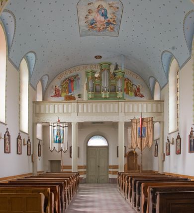 Zdjęcie nr 7: W prezbiterium do wysokości parapetów okiennych ściany malowane jednolicie w kolorze beżowym. Powyżej, na sklepieniu motyw gwiaździstego nieba z niewielkimi malowanymi złotem gwiazdkami na jasnobłękitnym tle. Ponad ołtarzem głównym gołębica Ducha Świętego na tle glorii promienistej tworzącej motyw dekoracyjnej rozety. Obramienia okienne zaznaczone geometrycznymi, złotymi lamówkami; rozglifienia okien białe. W podniebiu arkady tęczowej pas stylizowanego, symetrycznego ornamentu roślinnego w kolorach jasnoniebieskim, beżowym i jasnobrązowym. W osi uskrzydlona główka anielska. Na licu ściany tęczowej majuskułowy, złoty napis „NAPISANO JEST, ŻE DOM MÓJ JEST DOMEM MODLITWY”. W nawie głównej i transepcie ściany malowane jednolicie w kolorze beżowym. W południowym ramieniu transeptu, ponad ołtarzem motyw czteroliścia z wpisanym medalionem mieszczącym gorejące serce Jezusa, oplecione cierniową koroną i zwieńczone krzyżem. Na ścianie zachodniej, ponad chórem muzycznym dwie ustawione przeciwstawnie grupy muzykujących aniołów umieszczone na tle obłoków oraz majuskułowy, złoty napis „ŚPIEWAJCIE PANU W RADOŚCI”. Na artykułujących nawę filarach przechodzących w gurty sklepienne stylizowany, symetryczny ornament roślinny o przebiegu pasowym z wplecionymi motywami rozet z wpisanymi krzyżami równoramiennymi lub uskrzydlonymi główkami cherubinów. Ornament utrzymany w kolorach jasnozielonym, błękitnym, jasnobrązowym i beżowym. Na licu gurtu sklepiennego od strony zachodniej majuskułowy, złoty napis „NIECHAJ BĘDZIE POCHWALONY PRZENAJŚWIĘTSZY SAKRAMENT”. Sklepienie krzyżowe na przecięciu naw i ramionach transeptu oraz kolebkowe w nawie głównej malowane kolorem jasnobłękitnym w motyw złoconych gwiazd i rozetek. W wysklepkach, na przecięciu naw medaliony z popiersiami czterech ewangelistów. Od strony prezbiterium św. Mateusz ukazany jako dojrzały, ciemnowłosy mężczyzna piszący piórem w otwartej księdze, ubrany w czerwono-zielone szaty. Poniżej anioł – symbol św. Mateusza oraz banderola z napisem „ŚW(IĘTY) MATEUSZ”. Od południa medalion z popiersiem św. Jana ukazanego jako jasnowłosy młodzieniec, ubrany w zielono-fioletowe szaty, trzymający w ręku kielich. Poniżej orzeł o szeroko rozpostartych skrzydłach – symbol św. Jana oraz banderola z napisem „ŚW(IĘTY) JAN”. Od strony zachodniej medalion mieszczący popiersie św. Marka przedstawionego jako dojrzały mężczyzna o krótkich, ciemnych włosach i brodzie ubrany w żółto-niebieskie szaty. Święty trzyma w ręku zamkniętą księgę. Poniżej uskrzydlony lew – symbol św. Marka oraz banderola z napisem „ŚW(IĘTY) MAREK”. Od północy medalion z popiersiem św. Łukasza ukazanego jako dojrzały mężczyzna o krótkich, ciemnych włosach i brodzie trzymający w ręku obraz oraz laskę. Poniżej uskrzydlony byk oraz banderola z napisem „ŚW(IĘTY) ŁUKASZ”. W części centralnej tego przęsła medalion z czterema uskrzydlonymi główkami anielskimi. W centrum kolebkowego sklepienia ponad nawą główną pole w kształcie stojącego prostokąta zamkniętego łukiem półkolistym mieszczące przedstawienie Matki Boskiej Niepokalanie Poczętej na tle obłoków oraz aniołów i uskrzydlonych główek anielskich. Maria została przedstawiona jako młoda, jasnowłosa kobieta z rękami skrzyżowanymi na piersi, ubrana w jasnoróżową suknię oraz błękitny płaszcz. Pod jej stopami ukazano sierp księżyca. 