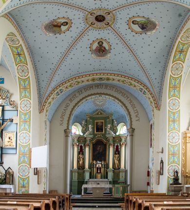 Zdjęcie nr 4: W prezbiterium do wysokości parapetów okiennych ściany malowane jednolicie w kolorze beżowym. Powyżej, na sklepieniu motyw gwiaździstego nieba z niewielkimi malowanymi złotem gwiazdkami na jasnobłękitnym tle. Ponad ołtarzem głównym gołębica Ducha Świętego na tle glorii promienistej tworzącej motyw dekoracyjnej rozety. Obramienia okienne zaznaczone geometrycznymi, złotymi lamówkami; rozglifienia okien białe. W podniebiu arkady tęczowej pas stylizowanego, symetrycznego ornamentu roślinnego w kolorach jasnoniebieskim, beżowym i jasnobrązowym. W osi uskrzydlona główka anielska. Na licu ściany tęczowej majuskułowy, złoty napis „NAPISANO JEST, ŻE DOM MÓJ JEST DOMEM MODLITWY”. W nawie głównej i transepcie ściany malowane jednolicie w kolorze beżowym. W południowym ramieniu transeptu, ponad ołtarzem motyw czteroliścia z wpisanym medalionem mieszczącym gorejące serce Jezusa, oplecione cierniową koroną i zwieńczone krzyżem. Na ścianie zachodniej, ponad chórem muzycznym dwie ustawione przeciwstawnie grupy muzykujących aniołów umieszczone na tle obłoków oraz majuskułowy, złoty napis „ŚPIEWAJCIE PANU W RADOŚCI”. Na artykułujących nawę filarach przechodzących w gurty sklepienne stylizowany, symetryczny ornament roślinny o przebiegu pasowym z wplecionymi motywami rozet z wpisanymi krzyżami równoramiennymi lub uskrzydlonymi główkami cherubinów. Ornament utrzymany w kolorach jasnozielonym, błękitnym, jasnobrązowym i beżowym. Na licu gurtu sklepiennego od strony zachodniej majuskułowy, złoty napis „NIECHAJ BĘDZIE POCHWALONY PRZENAJŚWIĘTSZY SAKRAMENT”. Sklepienie krzyżowe na przecięciu naw i ramionach transeptu oraz kolebkowe w nawie głównej malowane kolorem jasnobłękitnym w motyw złoconych gwiazd i rozetek. W wysklepkach, na przecięciu naw medaliony z popiersiami czterech ewangelistów. Od strony prezbiterium św. Mateusz ukazany jako dojrzały, ciemnowłosy mężczyzna piszący piórem w otwartej księdze, ubrany w czerwono-zielone szaty. Poniżej anioł – symbol św. Mateusza oraz banderola z napisem „ŚW(IĘTY) MATEUSZ”. Od południa medalion z popiersiem św. Jana ukazanego jako jasnowłosy młodzieniec, ubrany w zielono-fioletowe szaty, trzymający w ręku kielich. Poniżej orzeł o szeroko rozpostartych skrzydłach – symbol św. Jana oraz banderola z napisem „ŚW(IĘTY) JAN”. Od strony zachodniej medalion mieszczący popiersie św. Marka przedstawionego jako dojrzały mężczyzna o krótkich, ciemnych włosach i brodzie ubrany w żółto-niebieskie szaty. Święty trzyma w ręku zamkniętą księgę. Poniżej uskrzydlony lew – symbol św. Marka oraz banderola z napisem „ŚW(IĘTY) MAREK”. Od północy medalion z popiersiem św. Łukasza ukazanego jako dojrzały mężczyzna o krótkich, ciemnych włosach i brodzie trzymający w ręku obraz oraz laskę. Poniżej uskrzydlony byk oraz banderola z napisem „ŚW(IĘTY) ŁUKASZ”. W części centralnej tego przęsła medalion z czterema uskrzydlonymi główkami anielskimi. W centrum kolebkowego sklepienia ponad nawą główną pole w kształcie stojącego prostokąta zamkniętego łukiem półkolistym mieszczące przedstawienie Matki Boskiej Niepokalanie Poczętej na tle obłoków oraz aniołów i uskrzydlonych główek anielskich. Maria została przedstawiona jako młoda, jasnowłosa kobieta z rękami skrzyżowanymi na piersi, ubrana w jasnoróżową suknię oraz błękitny płaszcz. Pod jej stopami ukazano sierp księżyca. 