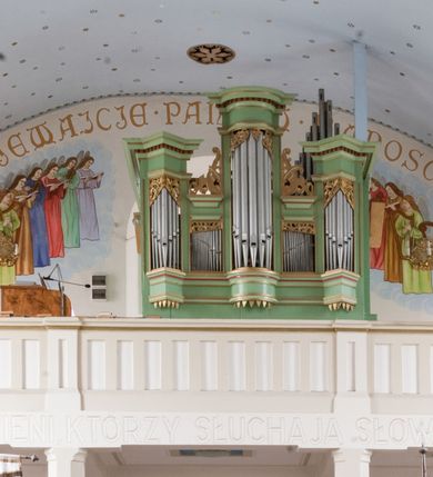 Zdjęcie nr 11: W prezbiterium do wysokości parapetów okiennych ściany malowane jednolicie w kolorze beżowym. Powyżej, na sklepieniu motyw gwiaździstego nieba z niewielkimi malowanymi złotem gwiazdkami na jasnobłękitnym tle. Ponad ołtarzem głównym gołębica Ducha Świętego na tle glorii promienistej tworzącej motyw dekoracyjnej rozety. Obramienia okienne zaznaczone geometrycznymi, złotymi lamówkami; rozglifienia okien białe. W podniebiu arkady tęczowej pas stylizowanego, symetrycznego ornamentu roślinnego w kolorach jasnoniebieskim, beżowym i jasnobrązowym. W osi uskrzydlona główka anielska. Na licu ściany tęczowej majuskułowy, złoty napis „NAPISANO JEST, ŻE DOM MÓJ JEST DOMEM MODLITWY”. W nawie głównej i transepcie ściany malowane jednolicie w kolorze beżowym. W południowym ramieniu transeptu, ponad ołtarzem motyw czteroliścia z wpisanym medalionem mieszczącym gorejące serce Jezusa, oplecione cierniową koroną i zwieńczone krzyżem. Na ścianie zachodniej, ponad chórem muzycznym dwie ustawione przeciwstawnie grupy muzykujących aniołów umieszczone na tle obłoków oraz majuskułowy, złoty napis „ŚPIEWAJCIE PANU W RADOŚCI”. Na artykułujących nawę filarach przechodzących w gurty sklepienne stylizowany, symetryczny ornament roślinny o przebiegu pasowym z wplecionymi motywami rozet z wpisanymi krzyżami równoramiennymi lub uskrzydlonymi główkami cherubinów. Ornament utrzymany w kolorach jasnozielonym, błękitnym, jasnobrązowym i beżowym. Na licu gurtu sklepiennego od strony zachodniej majuskułowy, złoty napis „NIECHAJ BĘDZIE POCHWALONY PRZENAJŚWIĘTSZY SAKRAMENT”. Sklepienie krzyżowe na przecięciu naw i ramionach transeptu oraz kolebkowe w nawie głównej malowane kolorem jasnobłękitnym w motyw złoconych gwiazd i rozetek. W wysklepkach, na przecięciu naw medaliony z popiersiami czterech ewangelistów. Od strony prezbiterium św. Mateusz ukazany jako dojrzały, ciemnowłosy mężczyzna piszący piórem w otwartej księdze, ubrany w czerwono-zielone szaty. Poniżej anioł – symbol św. Mateusza oraz banderola z napisem „ŚW(IĘTY) MATEUSZ”. Od południa medalion z popiersiem św. Jana ukazanego jako jasnowłosy młodzieniec, ubrany w zielono-fioletowe szaty, trzymający w ręku kielich. Poniżej orzeł o szeroko rozpostartych skrzydłach – symbol św. Jana oraz banderola z napisem „ŚW(IĘTY) JAN”. Od strony zachodniej medalion mieszczący popiersie św. Marka przedstawionego jako dojrzały mężczyzna o krótkich, ciemnych włosach i brodzie ubrany w żółto-niebieskie szaty. Święty trzyma w ręku zamkniętą księgę. Poniżej uskrzydlony lew – symbol św. Marka oraz banderola z napisem „ŚW(IĘTY) MAREK”. Od północy medalion z popiersiem św. Łukasza ukazanego jako dojrzały mężczyzna o krótkich, ciemnych włosach i brodzie trzymający w ręku obraz oraz laskę. Poniżej uskrzydlony byk oraz banderola z napisem „ŚW(IĘTY) ŁUKASZ”. W części centralnej tego przęsła medalion z czterema uskrzydlonymi główkami anielskimi. W centrum kolebkowego sklepienia ponad nawą główną pole w kształcie stojącego prostokąta zamkniętego łukiem półkolistym mieszczące przedstawienie Matki Boskiej Niepokalanie Poczętej na tle obłoków oraz aniołów i uskrzydlonych główek anielskich. Maria została przedstawiona jako młoda, jasnowłosa kobieta z rękami skrzyżowanymi na piersi, ubrana w jasnoróżową suknię oraz błękitny płaszcz. Pod jej stopami ukazano sierp księżyca. 