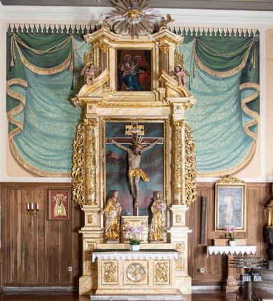 Zdjęcie nr 2: Nastawa ołtarzowa architektoniczna, dwukondygnacyjna, jednoosiowa z promienistą glorią w zwieńczeniu. Ołtarz prostopadłościenny, ustawiony na jednym stopniu, antependium zdobione dwiema prostokątnymi płycinami i środkową w kształcie kwadratu. Płyciny prostokątne wypełnione ornamentem akantowym, środkowa z medalionem, w polu którego umieszczony jest grecki krzyż o trójlistnie zakończonych ramionach, ukazany na tle czterech wiązek promieni. Tabernakulum w formie prostopadłościennej skrzynki z trzech stron flankowanej parami pilastrów hermowych, zamknięte profilowanym gzymsem. Drzwiczki w kształcie stojącego prostokąta, wypełnione rzeźbioną dekoracją manierystyczną. 

Dwukondygnacyjne retabulum ustawione jest na podwójnym cokole, ujęte parą pilastrów i ustawionych przed nimi kolumnami. Podpory wspierają wyłamane belkowanie i przerywany przyczółek, na którym usadzona jest para aniołków. Kolumny ustawione są na bazach, trzony bogato dekorowane winną latoroślą, ornamentem małżowinowo-chrząstkowym i główkami aniołków skierowanymi na wprost, kapitele kompozytowe. W polu głównym w prostokątnej, płytkiej wnęce umieszczony jest krucyfiks na tle malowanej Golgoty. Po bokach tabernakulum stoją ustawione rzeźby: św. Jana Ewangelisty po lewej stronie i Matki Boskiej po prawej stronie. Druga kondygnacja ustawiona na niskim cokole, ujęta parą zdwojonych pilastrów i parą kolumn podtrzymujących przerwane belkowanie, nad osią boczną ukośne, na którego szczycie usadzona jest para aniołków. W polu środkowym w ramie o kształcie stojącego prostokąta obraz św. Anny Samotrzeć. Na szczycie promienista gloria z półpostacią Boga Ojca na tle obłoków. Pierwsza i druga kondygnacja flankowane uszami z liści akantu. W partii fryzu dwie niewielkie główki aniołków, w pierwszej strefie cokołowej płyciny z liśćmi akantu, a w drugiej rozety. Struktura marmoryzowana w kolorze jasnobeżowym z czerwonym, brązowym i żółtym żyłkowaniem; ornamentyka i detale złocone oraz srebrzone.