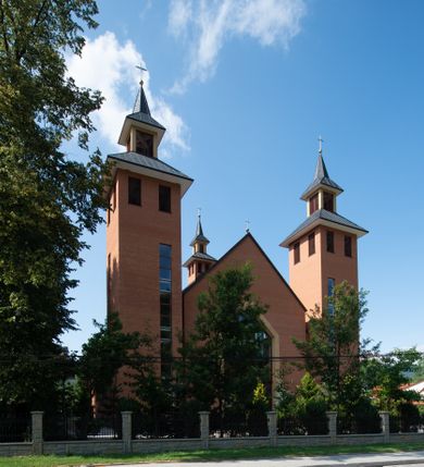 Zdjęcie nr 2: Kościół murowany, tynkowany, trójnawowy, bazylikowy. Prezbiterium prostokątne, zamknięte prosto, do północnej ściany przylega zakrystia. Nawa główna na planie prostokąta, o szerokości prezbiterium, nawy boczne węższe i krótsze. Na ich zakończeniu dostawione dwie wieże na planie kwadratu. Do południowej ściany korpusu nawowego przylega prostokątna kaplica, ustawiona pod kątem, otwarta do nawy prostokątnym prześwitem, z drzwiami w dolnej połowie i oknem w górnej. Prezbiterium i nawa główna sklepione kolebkowo z lunetami, nawy boczne kolebkowo. Nawa główna oddzielona od bocznych czterema parami kolumn. Parapet chóru muzycznego w zachodniej części nawy. Posadzka z płyt ceramicznych. 
Fasada zakończona trójkątnym szczytem. Na osi portal w formie stojącego prostokąta zamkniętego trójkątnie, w opasce tynkowej. W dolnej połowie dwuskrzydłowe drzwi, górna połowa przeszklona, podzielona na kwatery. Wieże pięciokondygnacyjne. Z wąskimi, przeszklonymi prześwitami przez cztery pierwsze kondygnacje na dwóch bokach. W piątej po dwa otwory dzwonne w formie stojącego prostokąta na każdej z czterech ścian. Wieże zwieńczone daszkami czterospadowymi oraz latarniami z otworami dzwonnymi, zakończonymi dachami wieżowymi z krzyżami. Pomiędzy fasadą a wieżami wąskie partie muru z przeszklonymi prześwitami. Na elewacjach bocznych nawy głównej, ponad dachami naw bocznych po trzy okna w formie wydłużonego leżącego prostokąta. Na elewacji północnej duże okno w formie leżącego prostokąta zamkniętego łukiem półkolistym. Kaplica południowa z wejściem na ścianie zachodniej, w formie drewnianego portalu w kształcie stojącego prostokąta, z dwuskrzydłowymi drzwiami, z kwadratowym oknem ponad. Na jej ścianie południowej dwa duże okna w kształcie leżącego prostokąta, na dwóch poziomach. Elewacja wschodnia zakończona trójkątnym szczytem. 
Nawa główna i prezbiterium nakryte dachami dwuspadowymi, nawy boczne pulpitowymi, kaplica dwuspadowym. Pomiędzy nawą a prezbiterium wieżyczka na sygnaturkę z latarnią, nakryta dachem wieżowym.  




