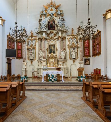 Zdjęcie nr 4: Kościół murowany, tynkowany, trójnawowy, bazylikowy. Prezbiterium prostokątne, zamknięte prosto, do północnej ściany przylega zakrystia. Nawa główna na planie prostokąta, o szerokości prezbiterium, nawy boczne węższe i krótsze. Na ich zakończeniu dostawione dwie wieże na planie kwadratu. Do południowej ściany korpusu nawowego przylega prostokątna kaplica, ustawiona pod kątem, otwarta do nawy prostokątnym prześwitem, z drzwiami w dolnej połowie i oknem w górnej. Prezbiterium i nawa główna sklepione kolebkowo z lunetami, nawy boczne kolebkowo. Nawa główna oddzielona od bocznych czterema parami kolumn. Parapet chóru muzycznego w zachodniej części nawy. Posadzka z płyt ceramicznych. 
Fasada zakończona trójkątnym szczytem. Na osi portal w formie stojącego prostokąta zamkniętego trójkątnie, w opasce tynkowej. W dolnej połowie dwuskrzydłowe drzwi, górna połowa przeszklona, podzielona na kwatery. Wieże pięciokondygnacyjne. Z wąskimi, przeszklonymi prześwitami przez cztery pierwsze kondygnacje na dwóch bokach. W piątej po dwa otwory dzwonne w formie stojącego prostokąta na każdej z czterech ścian. Wieże zwieńczone daszkami czterospadowymi oraz latarniami z otworami dzwonnymi, zakończonymi dachami wieżowymi z krzyżami. Pomiędzy fasadą a wieżami wąskie partie muru z przeszklonymi prześwitami. Na elewacjach bocznych nawy głównej, ponad dachami naw bocznych po trzy okna w formie wydłużonego leżącego prostokąta. Na elewacji północnej duże okno w formie leżącego prostokąta zamkniętego łukiem półkolistym. Kaplica południowa z wejściem na ścianie zachodniej, w formie drewnianego portalu w kształcie stojącego prostokąta, z dwuskrzydłowymi drzwiami, z kwadratowym oknem ponad. Na jej ścianie południowej dwa duże okna w kształcie leżącego prostokąta, na dwóch poziomach. Elewacja wschodnia zakończona trójkątnym szczytem. 
Nawa główna i prezbiterium nakryte dachami dwuspadowymi, nawy boczne pulpitowymi, kaplica dwuspadowym. Pomiędzy nawą a prezbiterium wieżyczka na sygnaturkę z latarnią, nakryta dachem wieżowym.  




