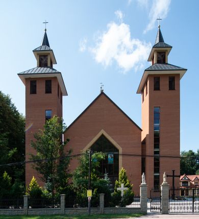 Zdjęcie nr 1: Kościół murowany, tynkowany, trójnawowy, bazylikowy. Prezbiterium prostokątne, zamknięte prosto, do północnej ściany przylega zakrystia. Nawa główna na planie prostokąta, o szerokości prezbiterium, nawy boczne węższe i krótsze. Na ich zakończeniu dostawione dwie wieże na planie kwadratu. Do południowej ściany korpusu nawowego przylega prostokątna kaplica, ustawiona pod kątem, otwarta do nawy prostokątnym prześwitem, z drzwiami w dolnej połowie i oknem w górnej. Prezbiterium i nawa główna sklepione kolebkowo z lunetami, nawy boczne kolebkowo. Nawa główna oddzielona od bocznych czterema parami kolumn. Parapet chóru muzycznego w zachodniej części nawy. Posadzka z płyt ceramicznych. 
Fasada zakończona trójkątnym szczytem. Na osi portal w formie stojącego prostokąta zamkniętego trójkątnie, w opasce tynkowej. W dolnej połowie dwuskrzydłowe drzwi, górna połowa przeszklona, podzielona na kwatery. Wieże pięciokondygnacyjne. Z wąskimi, przeszklonymi prześwitami przez cztery pierwsze kondygnacje na dwóch bokach. W piątej po dwa otwory dzwonne w formie stojącego prostokąta na każdej z czterech ścian. Wieże zwieńczone daszkami czterospadowymi oraz latarniami z otworami dzwonnymi, zakończonymi dachami wieżowymi z krzyżami. Pomiędzy fasadą a wieżami wąskie partie muru z przeszklonymi prześwitami. Na elewacjach bocznych nawy głównej, ponad dachami naw bocznych po trzy okna w formie wydłużonego leżącego prostokąta. Na elewacji północnej duże okno w formie leżącego prostokąta zamkniętego łukiem półkolistym. Kaplica południowa z wejściem na ścianie zachodniej, w formie drewnianego portalu w kształcie stojącego prostokąta, z dwuskrzydłowymi drzwiami, z kwadratowym oknem ponad. Na jej ścianie południowej dwa duże okna w kształcie leżącego prostokąta, na dwóch poziomach. Elewacja wschodnia zakończona trójkątnym szczytem. 
Nawa główna i prezbiterium nakryte dachami dwuspadowymi, nawy boczne pulpitowymi, kaplica dwuspadowym. Pomiędzy nawą a prezbiterium wieżyczka na sygnaturkę z latarnią, nakryta dachem wieżowym.  




