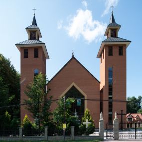 Zdjęcie nr 1: Kościół murowany, tynkowany, trójnawowy, bazylikowy. Prezbiterium prostokątne, zamknięte prosto, do północnej ściany przylega zakrystia. Nawa główna na planie prostokąta, o szerokości prezbiterium, nawy boczne węższe i krótsze. Na ich zakończeniu dostawione dwie wieże na planie kwadratu. Do południowej ściany korpusu nawowego przylega prostokątna kaplica, ustawiona pod kątem, otwarta do nawy prostokątnym prześwitem, z drzwiami w dolnej połowie i oknem w górnej. Prezbiterium i nawa główna sklepione kolebkowo z lunetami, nawy boczne kolebkowo. Nawa główna oddzielona od bocznych czterema parami kolumn. Parapet chóru muzycznego w zachodniej części nawy. Posadzka z płyt ceramicznych. 
Fasada zakończona trójkątnym szczytem. Na osi portal w formie stojącego prostokąta zamkniętego trójkątnie, w opasce tynkowej. W dolnej połowie dwuskrzydłowe drzwi, górna połowa przeszklona, podzielona na kwatery. Wieże pięciokondygnacyjne. Z wąskimi, przeszklonymi prześwitami przez cztery pierwsze kondygnacje na dwóch bokach. W piątej po dwa otwory dzwonne w formie stojącego prostokąta na każdej z czterech ścian. Wieże zwieńczone daszkami czterospadowymi oraz latarniami z otworami dzwonnymi, zakończonymi dachami wieżowymi z krzyżami. Pomiędzy fasadą a wieżami wąskie partie muru z przeszklonymi prześwitami. Na elewacjach bocznych nawy głównej, ponad dachami naw bocznych po trzy okna w formie wydłużonego leżącego prostokąta. Na elewacji północnej duże okno w formie leżącego prostokąta zamkniętego łukiem półkolistym. Kaplica południowa z wejściem na ścianie zachodniej, w formie drewnianego portalu w kształcie stojącego prostokąta, z dwuskrzydłowymi drzwiami, z kwadratowym oknem ponad. Na jej ścianie południowej dwa duże okna w kształcie leżącego prostokąta, na dwóch poziomach. Elewacja wschodnia zakończona trójkątnym szczytem. 
Nawa główna i prezbiterium nakryte dachami dwuspadowymi, nawy boczne pulpitowymi, kaplica dwuspadowym. Pomiędzy nawą a prezbiterium wieżyczka na sygnaturkę z latarnią, nakryta dachem wieżowym.  




