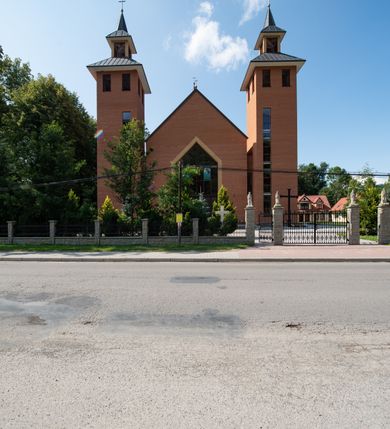 Zdjęcie nr 3: Kościół murowany, tynkowany, trójnawowy, bazylikowy. Prezbiterium prostokątne, zamknięte prosto, do północnej ściany przylega zakrystia. Nawa główna na planie prostokąta, o szerokości prezbiterium, nawy boczne węższe i krótsze. Na ich zakończeniu dostawione dwie wieże na planie kwadratu. Do południowej ściany korpusu nawowego przylega prostokątna kaplica, ustawiona pod kątem, otwarta do nawy prostokątnym prześwitem, z drzwiami w dolnej połowie i oknem w górnej. Prezbiterium i nawa główna sklepione kolebkowo z lunetami, nawy boczne kolebkowo. Nawa główna oddzielona od bocznych czterema parami kolumn. Parapet chóru muzycznego w zachodniej części nawy. Posadzka z płyt ceramicznych. 
Fasada zakończona trójkątnym szczytem. Na osi portal w formie stojącego prostokąta zamkniętego trójkątnie, w opasce tynkowej. W dolnej połowie dwuskrzydłowe drzwi, górna połowa przeszklona, podzielona na kwatery. Wieże pięciokondygnacyjne. Z wąskimi, przeszklonymi prześwitami przez cztery pierwsze kondygnacje na dwóch bokach. W piątej po dwa otwory dzwonne w formie stojącego prostokąta na każdej z czterech ścian. Wieże zwieńczone daszkami czterospadowymi oraz latarniami z otworami dzwonnymi, zakończonymi dachami wieżowymi z krzyżami. Pomiędzy fasadą a wieżami wąskie partie muru z przeszklonymi prześwitami. Na elewacjach bocznych nawy głównej, ponad dachami naw bocznych po trzy okna w formie wydłużonego leżącego prostokąta. Na elewacji północnej duże okno w formie leżącego prostokąta zamkniętego łukiem półkolistym. Kaplica południowa z wejściem na ścianie zachodniej, w formie drewnianego portalu w kształcie stojącego prostokąta, z dwuskrzydłowymi drzwiami, z kwadratowym oknem ponad. Na jej ścianie południowej dwa duże okna w kształcie leżącego prostokąta, na dwóch poziomach. Elewacja wschodnia zakończona trójkątnym szczytem. 
Nawa główna i prezbiterium nakryte dachami dwuspadowymi, nawy boczne pulpitowymi, kaplica dwuspadowym. Pomiędzy nawą a prezbiterium wieżyczka na sygnaturkę z latarnią, nakryta dachem wieżowym.  




