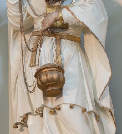 Zdjęcie nr 4: Rzeźba drążona z tyłu, ustawiona na niskiej podstawie. Postać zwrócona frontalnie, z opuszczona głową skierowaną w lewo. W lewej, ugiętej i uniesionej z przodu ręce trzyma złoconą kadzielnicę na łańcuszkach, które podtrzymuje również w uniesionej prawej ręce. Twarz ma pociągłą, o starczych rysach, dużych, migdałowatych i blisko siebie osadzonych oczach, dużym, prostym nosie i rozchylonych ustach, okoloną gęstą, silnie skręconą i uniesioną brodą oraz krótkimi włosami. Na głowie ma nakrycie kapłana. Ubrany jest w długą, białą suknię spodnią ze złoconymi lamówkami oraz białą suknię wierzchnią, sięgającą kolan, dekorowaną przy dolnej krawędzi złoconymi chwostami, przepasaną w talii złoconym pasem. Na ramiona ma narzucony biały płaszcz ze złoconymi lamówkami, opadający po bokach postaci, zaś na piersi ma złocony pektorał. Partie ciała polichromowane naturalistycznie.