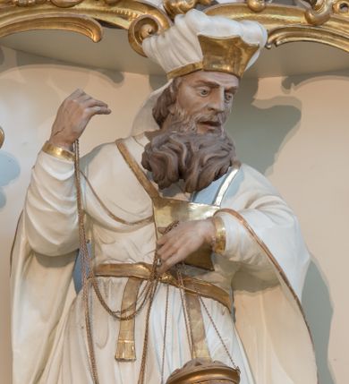 Zdjęcie nr 1: Rzeźba drążona z tyłu, ustawiona na niskiej podstawie. Postać zwrócona frontalnie, z opuszczona głową skierowaną w lewo. W lewej, ugiętej i uniesionej z przodu ręce trzyma złoconą kadzielnicę na łańcuszkach, które podtrzymuje również w uniesionej prawej ręce. Twarz ma pociągłą, o starczych rysach, dużych, migdałowatych i blisko siebie osadzonych oczach, dużym, prostym nosie i rozchylonych ustach, okoloną gęstą, silnie skręconą i uniesioną brodą oraz krótkimi włosami. Na głowie ma nakrycie kapłana. Ubrany jest w długą, białą suknię spodnią ze złoconymi lamówkami oraz białą suknię wierzchnią, sięgającą kolan, dekorowaną przy dolnej krawędzi złoconymi chwostami, przepasaną w talii złoconym pasem. Na ramiona ma narzucony biały płaszcz ze złoconymi lamówkami, opadający po bokach postaci, zaś na piersi ma złocony pektorał. Partie ciała polichromowane naturalistycznie.