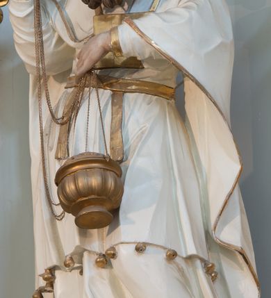 Zdjęcie nr 2: Rzeźba drążona z tyłu, ustawiona na niskiej podstawie. Postać zwrócona frontalnie, z opuszczona głową skierowaną w lewo. W lewej, ugiętej i uniesionej z przodu ręce trzyma złoconą kadzielnicę na łańcuszkach, które podtrzymuje również w uniesionej prawej ręce. Twarz ma pociągłą, o starczych rysach, dużych, migdałowatych i blisko siebie osadzonych oczach, dużym, prostym nosie i rozchylonych ustach, okoloną gęstą, silnie skręconą i uniesioną brodą oraz krótkimi włosami. Na głowie ma nakrycie kapłana. Ubrany jest w długą, białą suknię spodnią ze złoconymi lamówkami oraz białą suknię wierzchnią, sięgającą kolan, dekorowaną przy dolnej krawędzi złoconymi chwostami, przepasaną w talii złoconym pasem. Na ramiona ma narzucony biały płaszcz ze złoconymi lamówkami, opadający po bokach postaci, zaś na piersi ma złocony pektorał. Partie ciała polichromowane naturalistycznie.