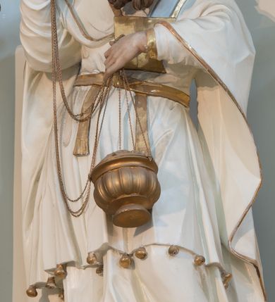 Zdjęcie nr 5: Rzeźba drążona z tyłu, ustawiona na niskiej podstawie. Postać zwrócona frontalnie, z opuszczona głową skierowaną w lewo. W lewej, ugiętej i uniesionej z przodu ręce trzyma złoconą kadzielnicę na łańcuszkach, które podtrzymuje również w uniesionej prawej ręce. Twarz ma pociągłą, o starczych rysach, dużych, migdałowatych i blisko siebie osadzonych oczach, dużym, prostym nosie i rozchylonych ustach, okoloną gęstą, silnie skręconą i uniesioną brodą oraz krótkimi włosami. Na głowie ma nakrycie kapłana. Ubrany jest w długą, białą suknię spodnią ze złoconymi lamówkami oraz białą suknię wierzchnią, sięgającą kolan, dekorowaną przy dolnej krawędzi złoconymi chwostami, przepasaną w talii złoconym pasem. Na ramiona ma narzucony biały płaszcz ze złoconymi lamówkami, opadający po bokach postaci, zaś na piersi ma złocony pektorał. Partie ciała polichromowane naturalistycznie.