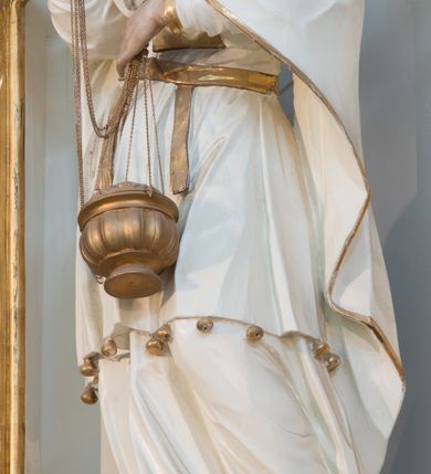 Zdjęcie nr 6: Rzeźba drążona z tyłu, ustawiona na niskiej podstawie. Postać zwrócona frontalnie, z opuszczona głową skierowaną w lewo. W lewej, ugiętej i uniesionej z przodu ręce trzyma złoconą kadzielnicę na łańcuszkach, które podtrzymuje również w uniesionej prawej ręce. Twarz ma pociągłą, o starczych rysach, dużych, migdałowatych i blisko siebie osadzonych oczach, dużym, prostym nosie i rozchylonych ustach, okoloną gęstą, silnie skręconą i uniesioną brodą oraz krótkimi włosami. Na głowie ma nakrycie kapłana. Ubrany jest w długą, białą suknię spodnią ze złoconymi lamówkami oraz białą suknię wierzchnią, sięgającą kolan, dekorowaną przy dolnej krawędzi złoconymi chwostami, przepasaną w talii złoconym pasem. Na ramiona ma narzucony biały płaszcz ze złoconymi lamówkami, opadający po bokach postaci, zaś na piersi ma złocony pektorał. Partie ciała polichromowane naturalistycznie.