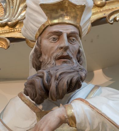 Zdjęcie nr 13: Rzeźba drążona z tyłu, ustawiona na niskiej podstawie. Postać zwrócona frontalnie, z opuszczona głową skierowaną w lewo. W lewej, ugiętej i uniesionej z przodu ręce trzyma złoconą kadzielnicę na łańcuszkach, które podtrzymuje również w uniesionej prawej ręce. Twarz ma pociągłą, o starczych rysach, dużych, migdałowatych i blisko siebie osadzonych oczach, dużym, prostym nosie i rozchylonych ustach, okoloną gęstą, silnie skręconą i uniesioną brodą oraz krótkimi włosami. Na głowie ma nakrycie kapłana. Ubrany jest w długą, białą suknię spodnią ze złoconymi lamówkami oraz białą suknię wierzchnią, sięgającą kolan, dekorowaną przy dolnej krawędzi złoconymi chwostami, przepasaną w talii złoconym pasem. Na ramiona ma narzucony biały płaszcz ze złoconymi lamówkami, opadający po bokach postaci, zaś na piersi ma złocony pektorał. Partie ciała polichromowane naturalistycznie.