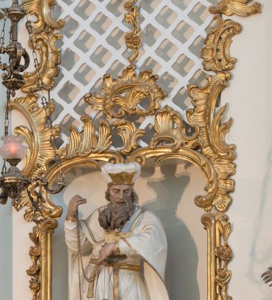 Zdjęcie nr 14: Rzeźba drążona z tyłu, ustawiona na niskiej podstawie. Postać zwrócona frontalnie, z opuszczona głową skierowaną w lewo. W lewej, ugiętej i uniesionej z przodu ręce trzyma złoconą kadzielnicę na łańcuszkach, które podtrzymuje również w uniesionej prawej ręce. Twarz ma pociągłą, o starczych rysach, dużych, migdałowatych i blisko siebie osadzonych oczach, dużym, prostym nosie i rozchylonych ustach, okoloną gęstą, silnie skręconą i uniesioną brodą oraz krótkimi włosami. Na głowie ma nakrycie kapłana. Ubrany jest w długą, białą suknię spodnią ze złoconymi lamówkami oraz białą suknię wierzchnią, sięgającą kolan, dekorowaną przy dolnej krawędzi złoconymi chwostami, przepasaną w talii złoconym pasem. Na ramiona ma narzucony biały płaszcz ze złoconymi lamówkami, opadający po bokach postaci, zaś na piersi ma złocony pektorał. Partie ciała polichromowane naturalistycznie.