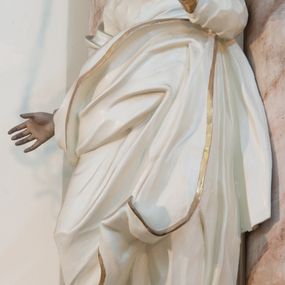 Zdjęcie nr 1: Rzeźba drążona z tyłu, ustawiona na niewielkiej podstawie. Święta zwrócona w trzech czwartych w lewo, w kontrapoście, z rękami rozłożonymi na boki. Twarz ma szczupłą, o starczych rysach, wyraźnie podkreślonej linii żuchwy, małych, migdałowatych oczach, prostym nosie i małych, rozchylonych ustach, okoloną włosami schowanymi pod welonem. Ubrana jest w długą, białą tunikę ze złoconą lamówką oraz biały płaszcz ze złoconą lamówką, zawinięty wokół prawego ramienia oraz wokół bioder, na głowie ma złocony welon z rozwianymi fragmentami tkaniny. Ciało polichromowane naturalistycznie. 