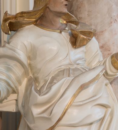 Zdjęcie nr 6: Rzeźba drążona z tyłu, ustawiona na niewielkiej podstawie. Święta zwrócona w trzech czwartych w lewo, w kontrapoście, z rękami rozłożonymi na boki. Twarz ma szczupłą, o starczych rysach, wyraźnie podkreślonej linii żuchwy, małych, migdałowatych oczach, prostym nosie i małych, rozchylonych ustach, okoloną włosami schowanymi pod welonem. Ubrana jest w długą, białą tunikę ze złoconą lamówką oraz biały płaszcz ze złoconą lamówką, zawinięty wokół prawego ramienia oraz wokół bioder, na głowie ma złocony welon z rozwianymi fragmentami tkaniny. Ciało polichromowane naturalistycznie. 