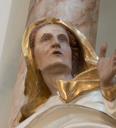 Zdjęcie nr 10: Rzeźba drążona z tyłu, ustawiona na niewielkiej podstawie. Święta zwrócona w trzech czwartych w lewo, w kontrapoście, z rękami rozłożonymi na boki. Twarz ma szczupłą, o starczych rysach, wyraźnie podkreślonej linii żuchwy, małych, migdałowatych oczach, prostym nosie i małych, rozchylonych ustach, okoloną włosami schowanymi pod welonem. Ubrana jest w długą, białą tunikę ze złoconą lamówką oraz biały płaszcz ze złoconą lamówką, zawinięty wokół prawego ramienia oraz wokół bioder, na głowie ma złocony welon z rozwianymi fragmentami tkaniny. Ciało polichromowane naturalistycznie. 