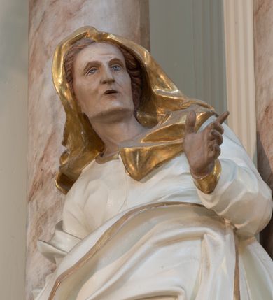 Zdjęcie nr 8: Rzeźba drążona z tyłu, ustawiona na niewielkiej podstawie. Święta zwrócona w trzech czwartych w lewo, w kontrapoście, z rękami rozłożonymi na boki. Twarz ma szczupłą, o starczych rysach, wyraźnie podkreślonej linii żuchwy, małych, migdałowatych oczach, prostym nosie i małych, rozchylonych ustach, okoloną włosami schowanymi pod welonem. Ubrana jest w długą, białą tunikę ze złoconą lamówką oraz biały płaszcz ze złoconą lamówką, zawinięty wokół prawego ramienia oraz wokół bioder, na głowie ma złocony welon z rozwianymi fragmentami tkaniny. Ciało polichromowane naturalistycznie. 