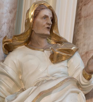 Zdjęcie nr 12: Rzeźba drążona z tyłu, ustawiona na niewielkiej podstawie. Święta zwrócona w trzech czwartych w lewo, w kontrapoście, z rękami rozłożonymi na boki. Twarz ma szczupłą, o starczych rysach, wyraźnie podkreślonej linii żuchwy, małych, migdałowatych oczach, prostym nosie i małych, rozchylonych ustach, okoloną włosami schowanymi pod welonem. Ubrana jest w długą, białą tunikę ze złoconą lamówką oraz biały płaszcz ze złoconą lamówką, zawinięty wokół prawego ramienia oraz wokół bioder, na głowie ma złocony welon z rozwianymi fragmentami tkaniny. Ciało polichromowane naturalistycznie. 