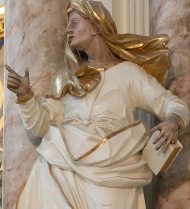 Zdjęcie nr 8: Rzeźba drążona z tyłu, ustawiona na niewielkiej podstawie. Święta zwrócona w trzech czwartych w prawo, w kontrapoście, lewa ręka przytrzymuje na boku księgę, prawa jest ugięta, uniesiona i wyciągnięta w bok. Twarz ma szczupłą, o wyraźnie podkreślonej linii żuchwy, migdałowatych, blisko siebie osadzonych oczach, dużym nosie i małych, rozchylonych ustach, okoloną włosami ukrytymi pod welonem. Ubrana jest w długą, białą tunikę ze złoconymi lamówkami, przylegającą do ciała w partii torsu oraz obfity, biały płaszcz ze złoconą lamówką zawinięty wokół bioder. Polichromia ciała naturalistyczna. 