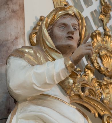 Zdjęcie nr 9: Rzeźba drążona z tyłu, ustawiona na niewielkiej podstawie. Święta zwrócona w trzech czwartych w prawo, w kontrapoście, lewa ręka przytrzymuje na boku księgę, prawa jest ugięta, uniesiona i wyciągnięta w bok. Twarz ma szczupłą, o wyraźnie podkreślonej linii żuchwy, migdałowatych, blisko siebie osadzonych oczach, dużym nosie i małych, rozchylonych ustach, okoloną włosami ukrytymi pod welonem. Ubrana jest w długą, białą tunikę ze złoconymi lamówkami, przylegającą do ciała w partii torsu oraz obfity, biały płaszcz ze złoconą lamówką zawinięty wokół bioder. Polichromia ciała naturalistyczna. 