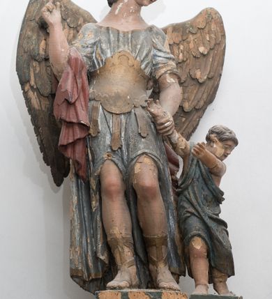 Zdjęcie nr 2: Figury anioła i dziecka ustawione są na niskich, osobnych podstawach. Anioł zwrócony jest frontalnie, z pochyloną głową skierowaną w lewo, w kontrapoście. Prawą rękę unosi w geście wskazującym, lewą trzyma za rączkę stojącego po jego lewej stronie chłopca. Twarz ma owalną, o niewielkich oczach skierowanych w dół, małym nosie i ustach, okoloną krótkimi, kręconymi włosami. Ma niewielką głowę osadzoną na długiej szyi. Ubrany jest w niebieską tunikę ze złoconymi lamówkami, z krótkimi rękawami, odsłaniającą nogi powyżej kolan. Wokół talii ma pancerz z trzema piórami, na prawym ramieniu ma zawieszony czerwony płaszcz, na nogach sandały. Skrzydła ma duże, złożone po bokach postaci. Po lewej stronie anioła stoi chłopiec zwrócony w trzech czwartych w lewo, w wykroku, z uniesionymi obiema rękami. Twarz ma owalną o niewielkich oczach skierowanych w dół, małym nosie i ustach, okoloną krótkimi włosami. Ubrany jest w zieloną szatę przewiązaną przez prawe ramię, sięgającą kolan. Polichromia ciała figur naturalistyczna.