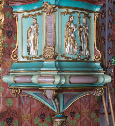 Zdjęcie nr 2: Ambona wisząca, przyścienna, z wejściem od zakrystii przez zaplecek. Kosz na planie czworoboku o ściętych narożach, o wypukłej ściance przedniej i wklęsłych bocznych, na profilowanym cokole oraz spłaszczonym wałku dekorowanym płycinami. U spodu kosza podwieszenie w formie stożka o wklęsłych bokach, ujętego w narożach karbowanymi wolutami, zakończonego szyszką ozdobioną rozetami. Kosz ujęty w narożach pilastrami, o trzonach w formie wolut dekorowanych rocaille&#039;em. Na ściankach rocaille&#039;owe płyciny z płaskorzeźbionymi przedstawieniami ewangelistów, na ściance bocznej lewej św. Łukasz z wołem, na przedniej – po prawej św. Jan z orłem, po lewej św. Mateusz z aniołem, na ściance bocznej prawej św. Marek z lwem. Parapet kosza w formie gzymsu. Zaplecek w kształcie prostokątnej ramy, opasującej drzwi. Baldachim na planie analogicznym względem kosza w formie podstawy ujętej fryzem zdobionym rozetami i gzymsem dekorowanym kimationem, z podwieszonym lambrekinem z chwostami. Na baldachimie cztery woluty podtrzymujące cokół, na którym stoi figura Chrystusa Dobrego Pasterza. Na podniebieniu baldachimu płaskorzeźbiona gołębica Ducha Świętego w promienistej glorii. Struktura ambony polichromowana w kolorze jasnoniebieskim, z niebieskimi, fioletowymi i czerwonymi detalami. Ornamentyka i profile złocone, detale srebrzone. 

