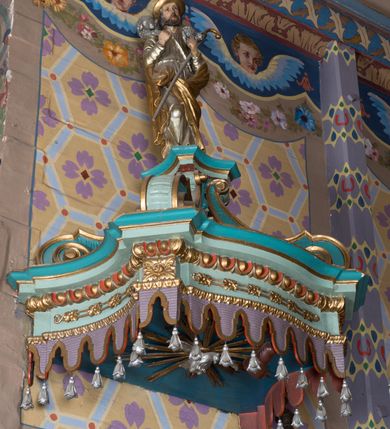 Zdjęcie nr 4: Ambona wisząca, przyścienna, z wejściem od zakrystii przez zaplecek. Kosz na planie czworoboku o ściętych narożach, o wypukłej ściance przedniej i wklęsłych bocznych, na profilowanym cokole oraz spłaszczonym wałku dekorowanym płycinami. U spodu kosza podwieszenie w formie stożka o wklęsłych bokach, ujętego w narożach karbowanymi wolutami, zakończonego szyszką ozdobioną rozetami. Kosz ujęty w narożach pilastrami, o trzonach w formie wolut dekorowanych rocaille'em. Na ściankach rocaille'owe płyciny z płaskorzeźbionymi przedstawieniami ewangelistów, na ściance bocznej lewej św. Łukasz z wołem, na przedniej – po prawej św. Jan z orłem, po lewej św. Mateusz z aniołem, na ściance bocznej prawej św. Marek z lwem. Parapet kosza w formie gzymsu. Zaplecek w kształcie prostokątnej ramy, opasującej drzwi. Baldachim na planie analogicznym względem kosza w formie podstawy ujętej fryzem zdobionym rozetami i gzymsem dekorowanym kimationem, z podwieszonym lambrekinem z chwostami. Na baldachimie cztery woluty podtrzymujące cokół, na którym stoi figura Chrystusa Dobrego Pasterza. Na podniebieniu baldachimu płaskorzeźbiona gołębica Ducha Świętego w promienistej glorii. Struktura ambony polichromowana w kolorze jasnoniebieskim, z niebieskimi, fioletowymi i czerwonymi detalami. Ornamentyka i profile złocone, detale srebrzone. 

