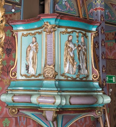 Zdjęcie nr 1: Ambona wisząca, przyścienna, z wejściem od zakrystii przez zaplecek. Kosz na planie czworoboku o ściętych narożach, o wypukłej ściance przedniej i wklęsłych bocznych, na profilowanym cokole oraz spłaszczonym wałku dekorowanym płycinami. U spodu kosza podwieszenie w formie stożka o wklęsłych bokach, ujętego w narożach karbowanymi wolutami, zakończonego szyszką ozdobioną rozetami. Kosz ujęty w narożach pilastrami, o trzonach w formie wolut dekorowanych rocaille'em. Na ściankach rocaille'owe płyciny z płaskorzeźbionymi przedstawieniami ewangelistów, na ściance bocznej lewej św. Łukasz z wołem, na przedniej – po prawej św. Jan z orłem, po lewej św. Mateusz z aniołem, na ściance bocznej prawej św. Marek z lwem. Parapet kosza w formie gzymsu. Zaplecek w kształcie prostokątnej ramy, opasującej drzwi. Baldachim na planie analogicznym względem kosza w formie podstawy ujętej fryzem zdobionym rozetami i gzymsem dekorowanym kimationem, z podwieszonym lambrekinem z chwostami. Na baldachimie cztery woluty podtrzymujące cokół, na którym stoi figura Chrystusa Dobrego Pasterza. Na podniebieniu baldachimu płaskorzeźbiona gołębica Ducha Świętego w promienistej glorii. Struktura ambony polichromowana w kolorze jasnoniebieskim, z niebieskimi, fioletowymi i czerwonymi detalami. Ornamentyka i profile złocone, detale srebrzone. 

