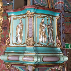 Zdjęcie nr 1: Ambona wisząca, przyścienna, z wejściem od zakrystii przez zaplecek. Kosz na planie czworoboku o ściętych narożach, o wypukłej ściance przedniej i wklęsłych bocznych, na profilowanym cokole oraz spłaszczonym wałku dekorowanym płycinami. U spodu kosza podwieszenie w formie stożka o wklęsłych bokach, ujętego w narożach karbowanymi wolutami, zakończonego szyszką ozdobioną rozetami. Kosz ujęty w narożach pilastrami, o trzonach w formie wolut dekorowanych rocaille'em. Na ściankach rocaille'owe płyciny z płaskorzeźbionymi przedstawieniami ewangelistów, na ściance bocznej lewej św. Łukasz z wołem, na przedniej – po prawej św. Jan z orłem, po lewej św. Mateusz z aniołem, na ściance bocznej prawej św. Marek z lwem. Parapet kosza w formie gzymsu. Zaplecek w kształcie prostokątnej ramy, opasującej drzwi. Baldachim na planie analogicznym względem kosza w formie podstawy ujętej fryzem zdobionym rozetami i gzymsem dekorowanym kimationem, z podwieszonym lambrekinem z chwostami. Na baldachimie cztery woluty podtrzymujące cokół, na którym stoi figura Chrystusa Dobrego Pasterza. Na podniebieniu baldachimu płaskorzeźbiona gołębica Ducha Świętego w promienistej glorii. Struktura ambony polichromowana w kolorze jasnoniebieskim, z niebieskimi, fioletowymi i czerwonymi detalami. Ornamentyka i profile złocone, detale srebrzone. 

