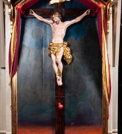 Zdjęcie nr 1: Rzeźbiony krucyfiks, zawieszony na prostym krzyżu na tle obrazu z przedstawieniem zaćmienia słońca w górnej części i widokiem Jerozolimy za wzgórzami w dolnej, w niszy zamkniętej od góry daszkiem dwuspadowym. W centrum rzeźbiona figura Chrystusa Ukrzyżowanego, z rozłożonymi, nieznacznie ugiętymi rękami przybitymi gwoździami, ze stopami złożonymi razem, prawa na lewej, przybitymi jednym gwoździem; lewe kolano ugięte, wysunięte przed prawe. Prawe biodro lekko wygięte, ciało tworzy mało wydatny łuk. Na torsie widoczna rana w prawym boku. Głowa opada na prawe ramię, twarz owalna, oczy zamknięte, brwi lekko uniesione, nos prosty, widoczne krótkie wąsy i dzieląca się w dwa pukle broda. Kręcone włosy opadają na kark i prawe ramię. Zarost i włosy jasnobrązowe. Na głowie masywna, złocona korona cierniowa. Biodra przewiązane złoconym, krótkim perizonium związanym na lewym biodrze, układającym się w drobne, poprzeczne fałdy. Koniec perizonium powiewa przy lewym udzie. Chrystus wisi na drewnianym, ciemnobrązowym krzyżu o długiej belce pionowej. W jej górnej części titulus z napisem „INRI”. 